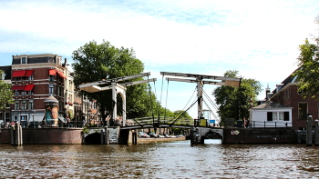 Магере-Брюг / Магере-Брюг (или «Тощий мост») находится в самом центре Амстердама, перебросив свои изящные, но куцые крылья меж двух берегов реки Амстел. С одной стороны от моста лежит улица Керкстраат, а с другой, что легко запомнить, Ньиве-Керкстраат. И вот тут самое время рассказать о легенде возникновения названия моста. Говорят, давным давно на разных берегах этого канала жили две сестры, которым, для того чтобы встретиться, необходимо было делать немалый крюк до ближайшего моста. Покумекав, они решили построить мост для себя: очень узкий и небольшой. Так и появилось его название — «тощий». Вторая легенда гласит, что Магере (с голландского «тощий») звалось семейство, которое возвело этот самый мост. В наследство от семьи-владельца переправе досталось и такое имя.Сегодня раздвижной деревянный мост белого цвета — важная достопримечательность города. Каждый май, когда в Европе отмечают День Победы, на нем дают праздничный концерт. Который удостаивает своим вниманием сама королева Беатрикс. А еще мост «засветился» в знаменитой бондиане: здесь снималась одна из сцен фильма о Джеймсе Бонде 1971 г. «Бриллианты навсегда».Как уже говорилось выше, этот мост раздвижной.