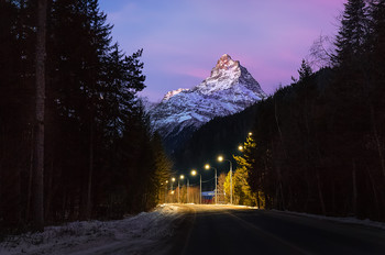 Гора Белалакая / Ранняя заря. Ещё темно, горят фонари. Но рассветное небо уже окрасилось розовым оттенком. Гора, будто из иллюстрации сказки, встречает гостей курорта. 
Этот вид дороги с вершиной – одна из визитных карточек Домбая. Белалака́я («Полосатая скала») — вершина западной части главного хребта Большого Кавказа. Высота — 3861 метр.
21 декабря, 2019 г. 
Фотопроект «Кавказ без границ».